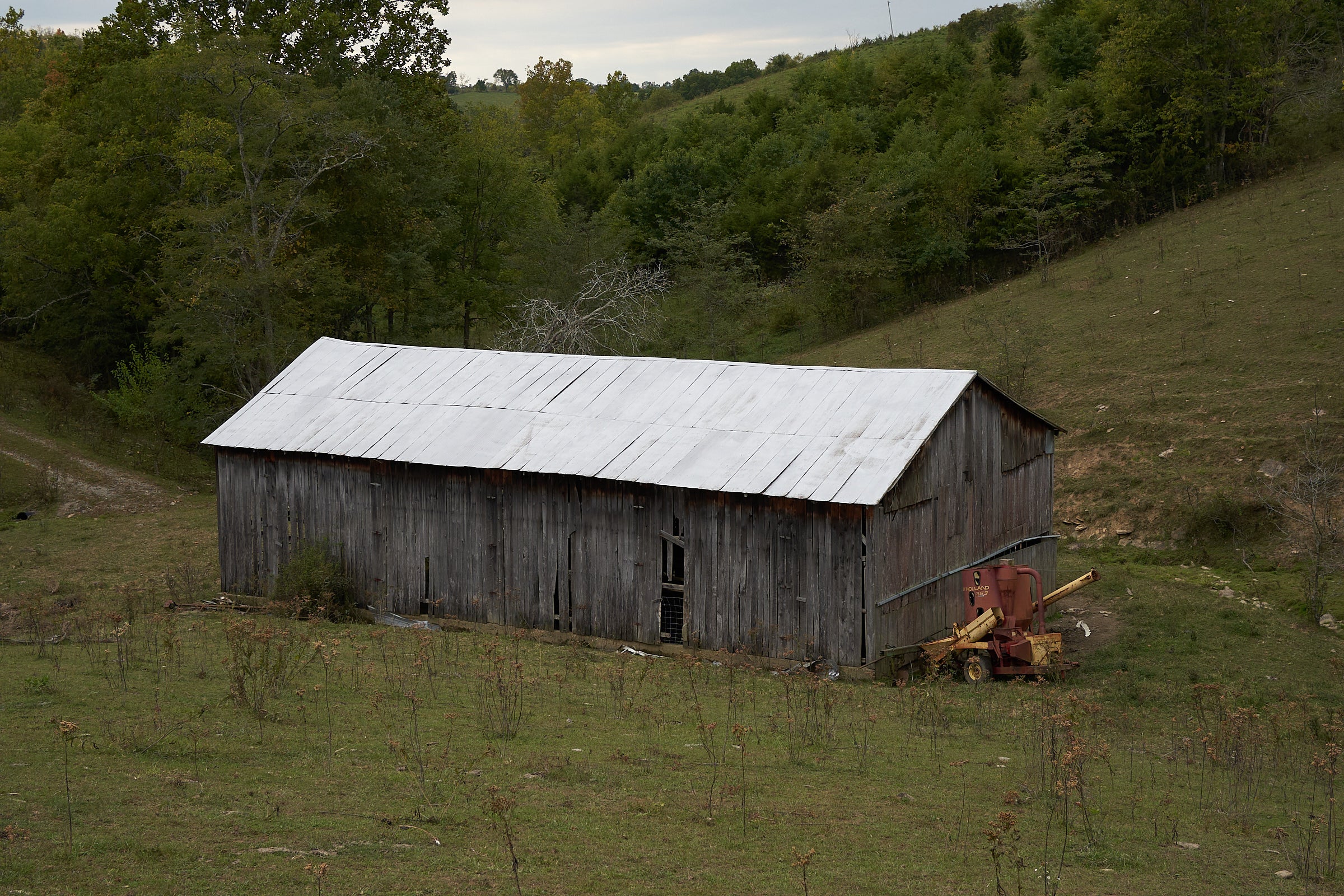 Old wooden barn in a rural landscape with trees and hills in the background.